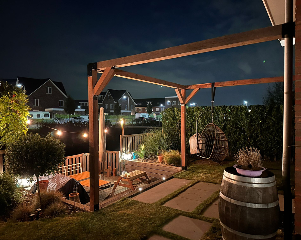 Houten pergola (12x12 cm) aan de muur gemonteerd, sfeervol verlicht in een tuin bij nacht. Douglas constructie met metalen koppelstukken voor een luxe buitenruimte