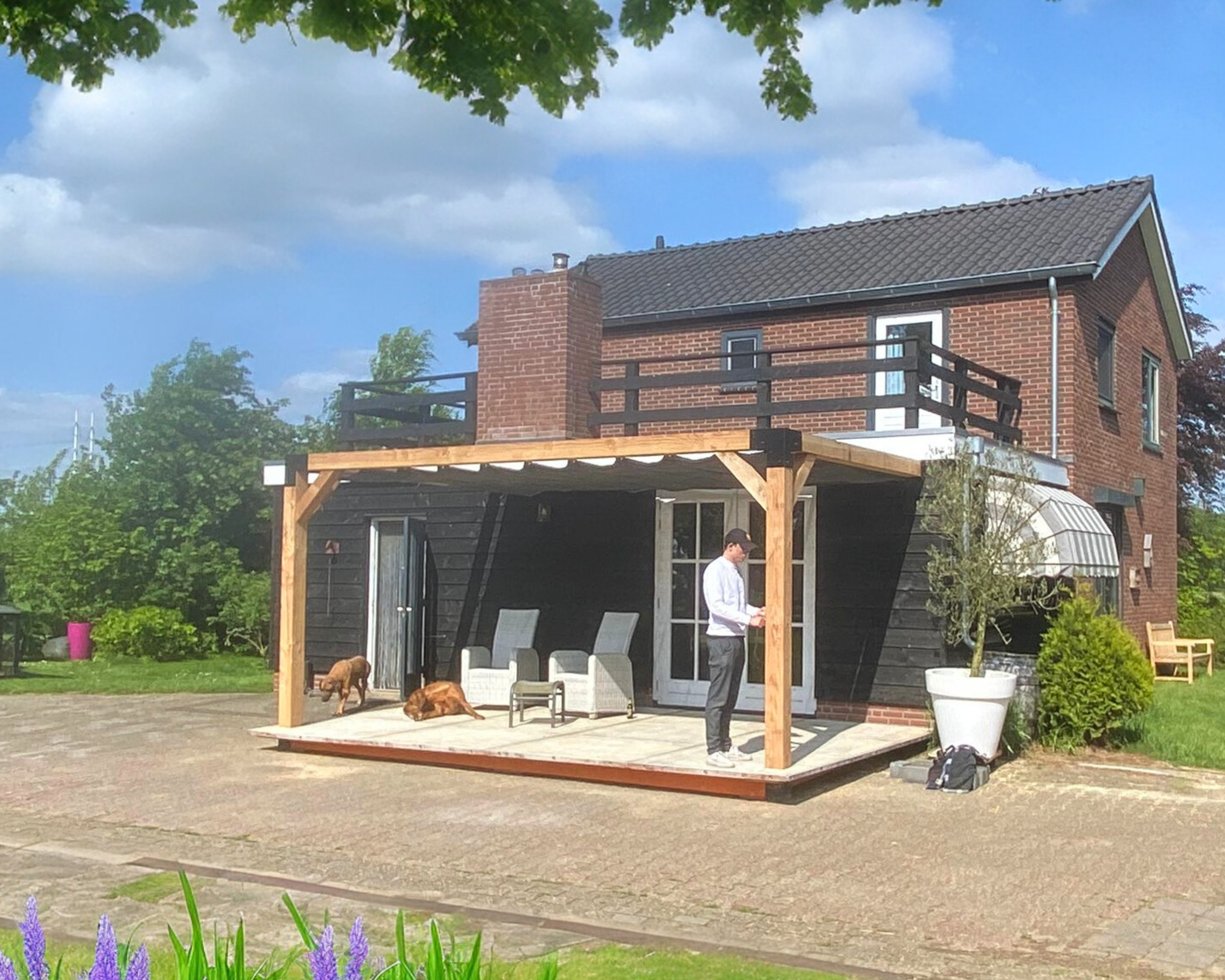 Houten muurpergola (15x15 cm) met wit harmonicadoek, gemonteerd aan een landelijke woning. Luxe terrasoverkapping van Douglas hout, perfect voor een sfeervolle buitenruimte